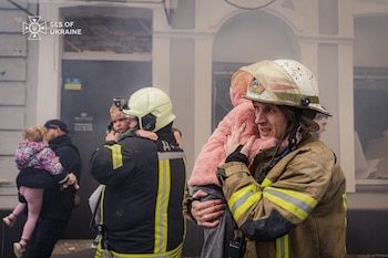 Primer plano de un bombero ucraniano con casco sucio, llevando a un niño envuelto en una prenda rosa. Al fondo, otro rescatista con un niño y humo