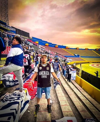 Joven con camiseta negra y gorra posa en una grada de estadio de fútbol azul y amarillo, lleno de gente y banderas, bajo un cielo naranja al atardecer