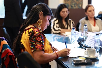 Mujer de perfil con cabello oscuro, blusa tradicional amarilla y de colores, aretes de cuentas, escribiendo con bolígrafo azul en una mesa con documentos