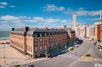 Vista aérea de un gran edificio de ladrillo rojo y techo oscuro con ventanas blancas, ubicado junto a una playa, con calles y rascacielos urbanos bajo un cielo azul nublado