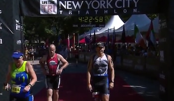 Tres deportistas cruzan la línea de meta de un triatlón. Un arco indica 'New York City Triathlon' y un reloj digital marca 4:22:58