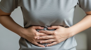 Vista cercana del torso de una persona con camiseta gris, sosteniendo su abdomen con ambas manos, los dedos ligeramente apretados.