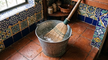 Balde metálico con agua, sal y una escoba de fibra sumergida en una cocina con azulejos decorados y suelo de barro.