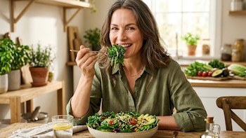 Mulher adulta feliz comendo uma grande salada verde em uma cozinha iluminada. Ele está segurando um garfo com legumes. A salada contém abacate, tomate e sementes