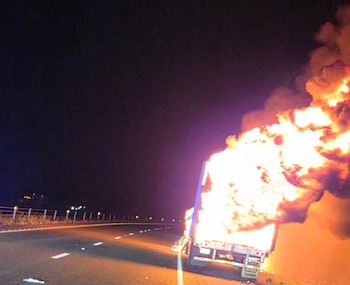 Vista trasera de un camión envuelto en grandes llamas anaranjadas y humo oscuro en el arcén de una carretera oscura por la noche