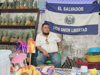 Hombre y mujer asiáticos sonriendo en primer plano, la mujer sostiene un letrero con forma de piña que dice "Feria de la Piña 2026 Santa María Ostuma"