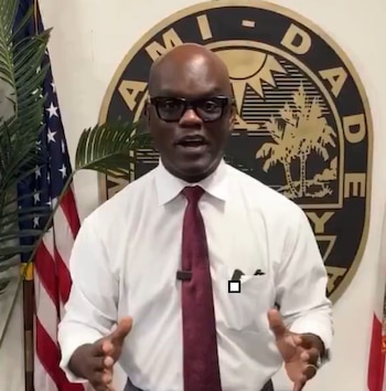 Un hombre con camisa blanca y corbata gesticula frente al sello del condado de Miami-Dade y la bandera de Estados Unidos