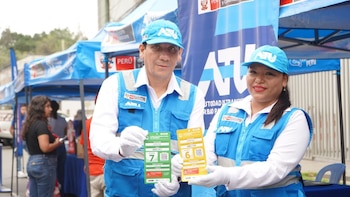 Dos personas con chalecos y gorros azules sostienen dos tarjetas impresas, una verde y otra amarilla, frente a un módulo al aire libre