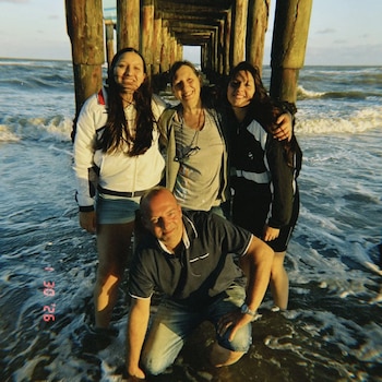Un hombre arrodillado y tres mujeres de pie posan en la orilla del mar debajo de los pilares de madera de un muelle