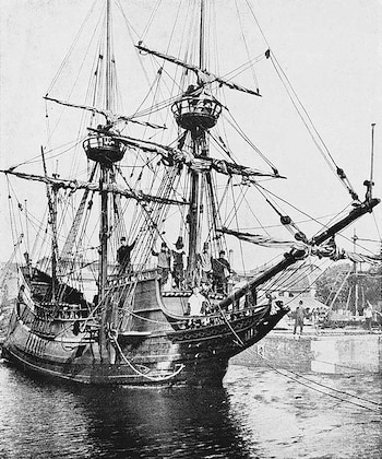Fotografía en blanco y negro de un barco de vela de tres mástiles amarrado en un muelle, con varias personas a bordo y en la orilla