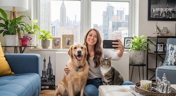 Una mujer sonriente sentada entre un perro labrador dorado y un gato atigrado, tomando una selfie con su teléfono frente a una ventana con vista a la ciudad de Nueva York.