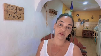 Una mujer en primer plano delante de una pared blanca con un cartel de madera y una cocina al fondo.