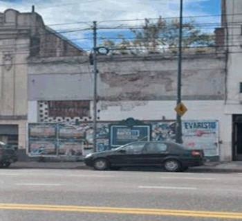 Coche sedán negro estacionado en la calle junto a un edificio antiguo de dos pisos con fachada desgastada, postes de luz y cables visibles