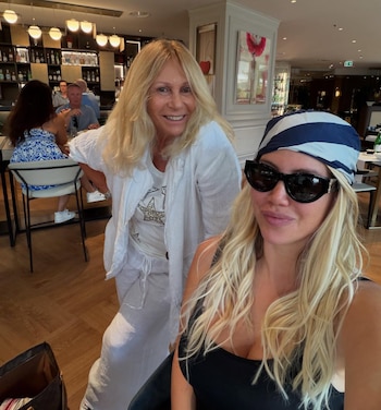Una mujer de pie de cabello rubio, vestida de blanco, y otra mujer sentada de cabello rubio, con gafas de sol y un pañuelo a rayas