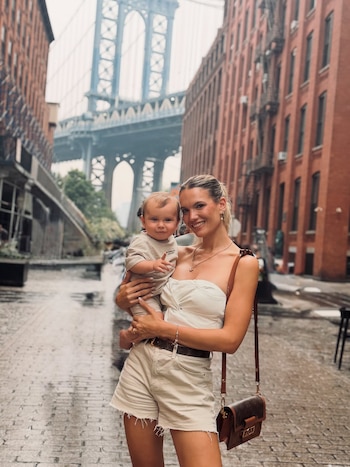Una mujer joven con un bebé en brazos posa sonriendo en una calle. Detrás, hay edificios de ladrillo y el puente de Manhattan