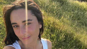 Camila Arena, mujer de cabello oscuro y camiseta blanca, sonríe levemente con fuerte luz solar. Fondo de hierba alta verde borrosa