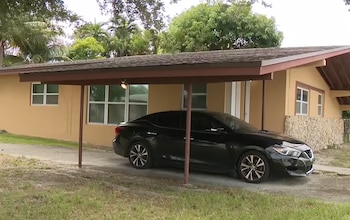 Una casa de un piso de color beis con garaje techado, un automóvil negro estacionado y árboles al fondo