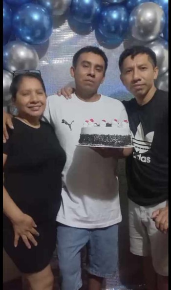 Tres personas posan para una foto. El hombre del centro, con camiseta blanca, sostiene un pastel. Una mujer y otro hombre lo acompañan sonriendo