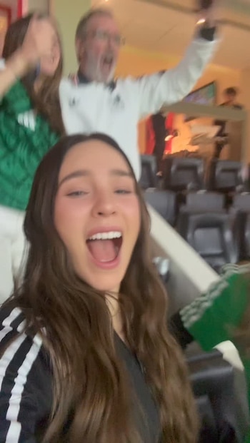 Primer plano de mujer con cabello castaño largo y boca abierta. Detrás, dos personas borrosas, una con camiseta verde y otra con chaqueta blanca, también celebran