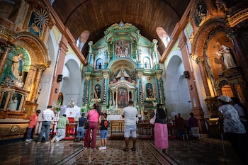 Vista interior de una iglesia con un altar mayor barroco y ricamente decorado en tonos verde y dorado. Varias personas observan el altar, algunas arrodilladas