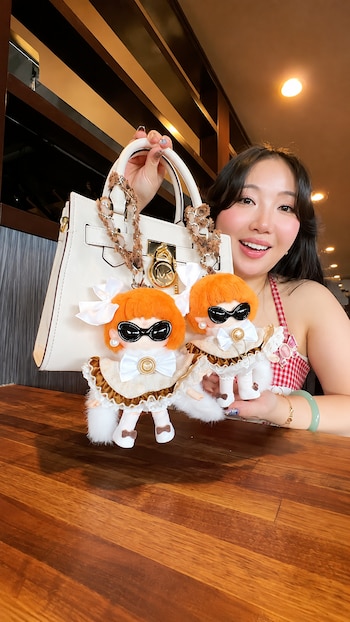 Mujer sonriente, bolso de mano blanco, dos muñecas de llavero con cabello naranja y gafas, mesa de madera, fondo oscuro
