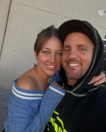 Primer plano de una mujer de cabello castaño y un hombre con barba y gorra, sonriendo mientras se abrazan, con una pared gris de fondo