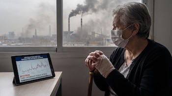 Mujer mayor con mascarilla, muñeca vendada, y bastón. Ventana con vista a fábricas que emiten humo. Tableta en mesa con gráfico y noticia sobre contaminación del aire.