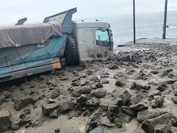 Vista de un camión de carga semivolcado en un camino cubierto de lodo y rocas. Al fondo, se observa el mar y un cielo nublado con postes eléctricos