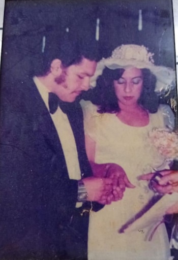 Foto vintage de boda: un hombre con traje oscuro y pajarita y una mujer con vestido blanco y sombrero se miran las manos entrelazadas