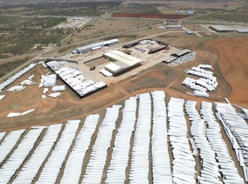 Vista aérea de un vasto campo con miles de palas de turbinas eólicas blancas, organizadas en filas, junto a edificios industriales y caminos