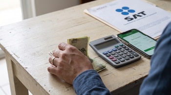 Una mano cuenta billetes de pesos mexicanos sobre una mesa de madera clara. Junto a la mano hay una calculadora, un teléfono móvil y un documento con el logo del SAT.