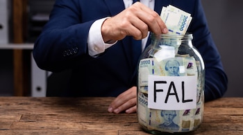 Mano de hombre con traje introduce billetes de 20.000 pesos argentinos en frasco de vidrio con etiqueta que dice FAL, sobre mesa de madera.