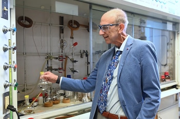 Hombre con gafas de seguridad y chaqueta azul sostiene un matraz con líquido transparente en un laboratorio. Se ve un panel de control con perillas y equipo de vidrio