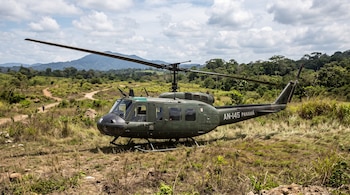 Helicóptero verde con la inscripción AN-145 Panamá posado sobre hierba alta en un campo con colinas y árboles al fondo.