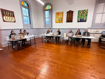 Nueve personas sentadas detrás de tres mesas largas con cajas de votación en un salón con piso de madera y paredes blancas, con ventanas y cuadros abstractos