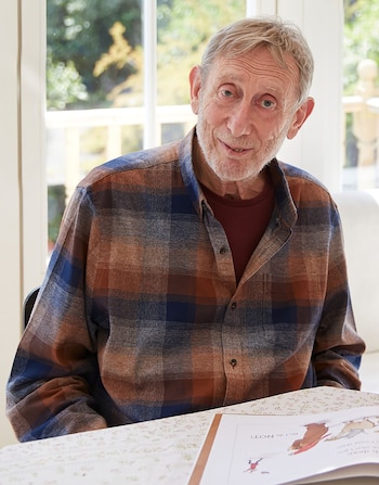 Michael Rosen, hombre mayor con cabello y barba gris, camisa a cuadros marrón y azul. Sonríe a la cámara, sentado frente a una ventana, con un libro abierto sobre la mesa