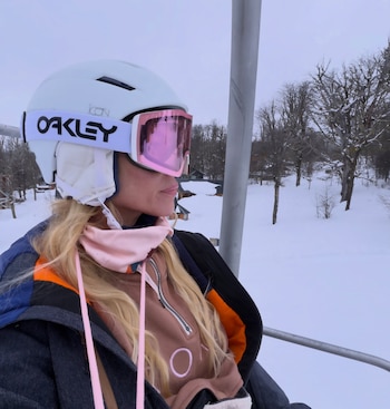 Primer plano de una mujer rubia con casco blanco y antiparras rosadas en una telesilla. De fondo, paisaje nevado con árboles sin hojas
