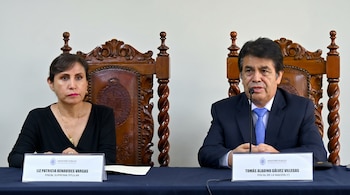 Dos personas, una mujer y un hombre, sentadas en una mesa con micrófonos. Llevan sillas de madera ornamentadas y sus nombres están en carteles.