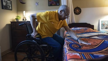 Max Gomez, um idoso de camisa amarela e óculos, apoia-se numa cadeira de rodas Invacare para ser transferido para a cama com o conforto do conforto.