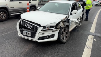 Automóvil Audi blanco con frente destrozado, rueda delantera derecha doblada, dos agentes de policía y otros vehículos en una calle de asfalto