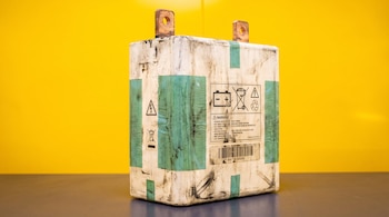 Batería rectangular blanca con suciedad y cinta verde, dos terminales de cobre, etiquetas de advertencia, código de barras, sobre superficie gris y fondo amarillo.