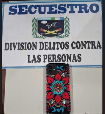 Teléfono celular con funda multicolor sobre una superficie blanca, delante de un cartel que dice "SECUESTRO" y "DIVISIÓN DELITOS CONTRA LAS PERSONAS"