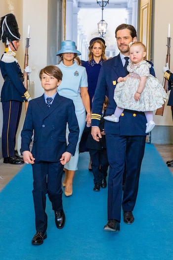 Miembros de la realeza, incluyendo un hombre con un bebé en brazos, caminan por un pasillo con alfombra azul, flanqueados por guardias