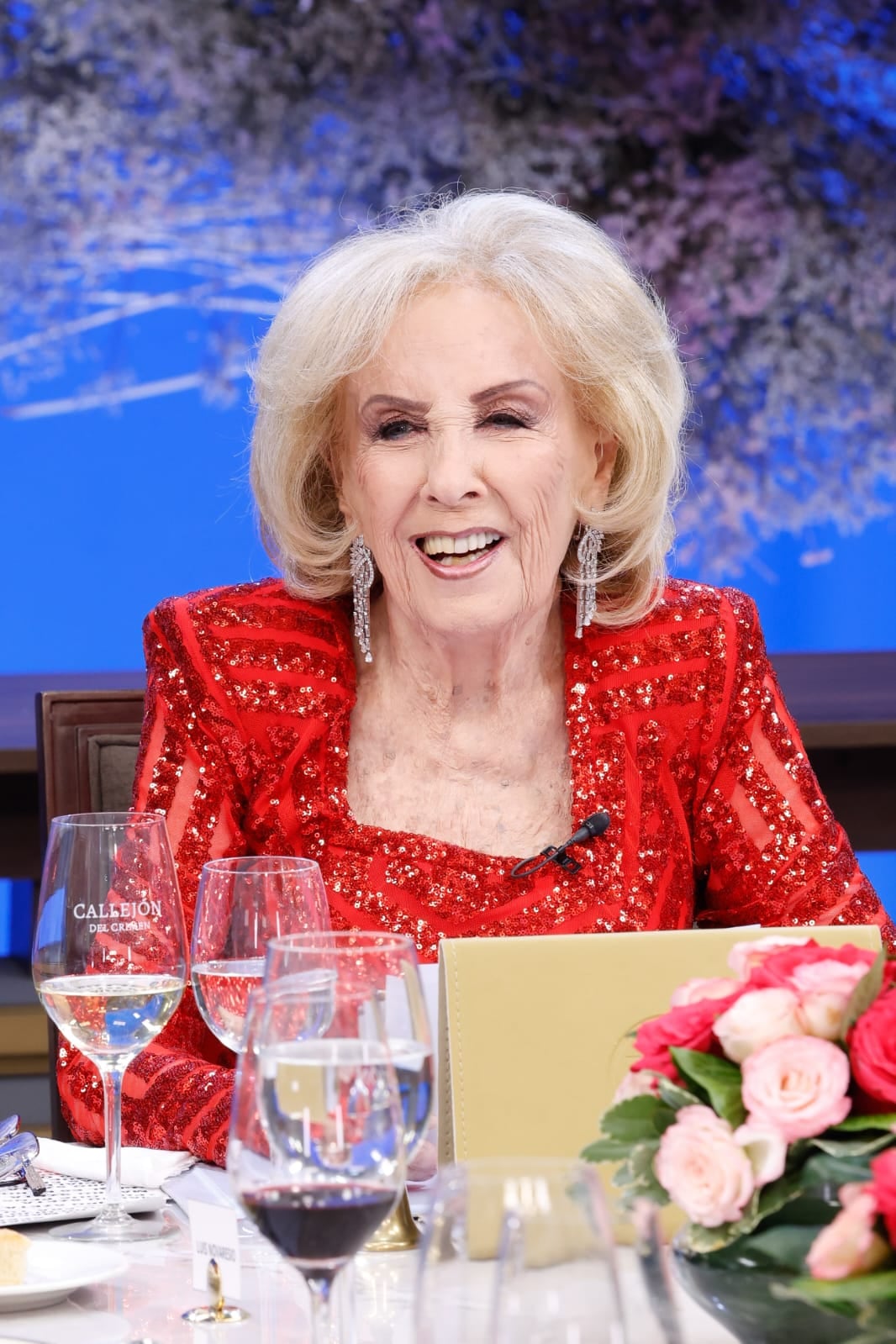 Mirtha Legrand, la icónica conductora, sonríe ampliamente mientras luce un elegante conjunto rojo de lentejuelas y largos aros, sentada a una mesa adornada con copas y un arreglo floral, irradiando su clásico glamour televisivo.