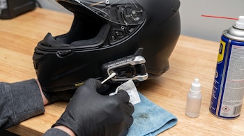 Una persona con guantes negros limpia un intercomunicador Cardo Packtalk Bold adherido a un casco de motocicleta negro, con un hisopo, una toalla y un líquido limpiador.