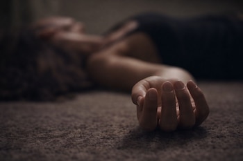 Una mano de mujer, con los dedos ligeramente curvados, en primer plano y en foco sobre una alfombra marrón. Detrás, el cuerpo de una mujer con ropa oscura aparece desenfocado.