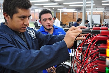 Primer plano de un joven en uniforme azul conectando cables rojos y negros a un panel electrónico, con otro joven observando detrás en un laboratorio