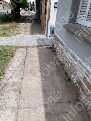 Vereda de cemento con grietas y desniveles, áreas con pasto, pared de ladrillos y calle con árboles y edificios en el fondo