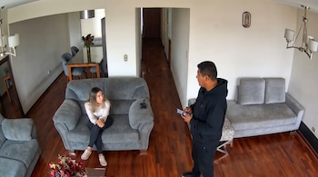 Dos personas, un hombre y una mujer, están en una sala de estar con sofás grises, piso de madera y una mesa de comedor al fondo.