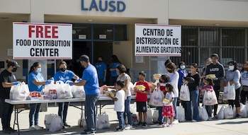 Voluntarios y personal escolar enmascarados entregan bolsas de alimentos a familias con niños. Al fondo, una escuela con el letrero LAUSD y señalización de "centro de distribución".